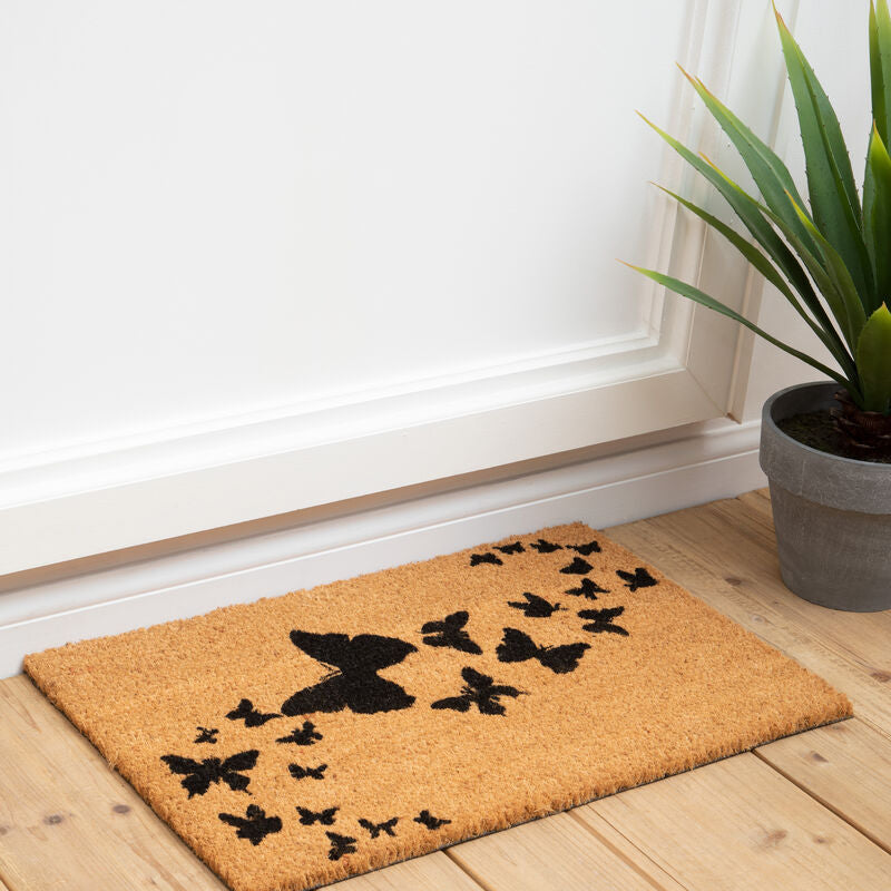 Doormat with butterfly design on a wooden floor next to a plant