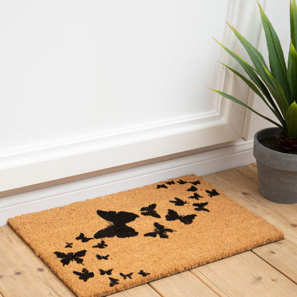 Doormat with butterfly design on a wooden floor next to a plant