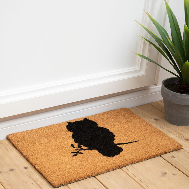 Doormat with owl design on a wooden floor next to a plant