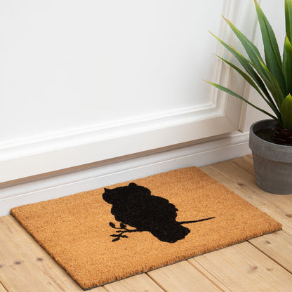 Doormat with owl design on a wooden floor next to a plant