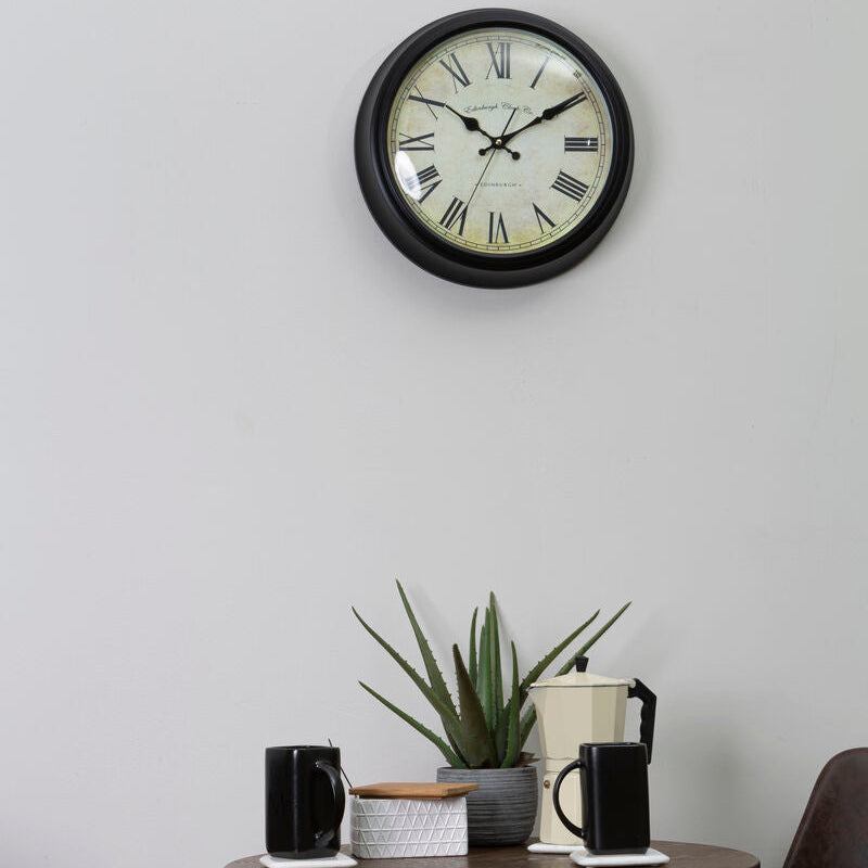 Wall clock on a light gray wall above a small table with a plant and coffee cups.