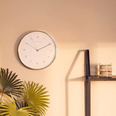 Round wall clock on a beige wall with a plant and shelf in the foreground