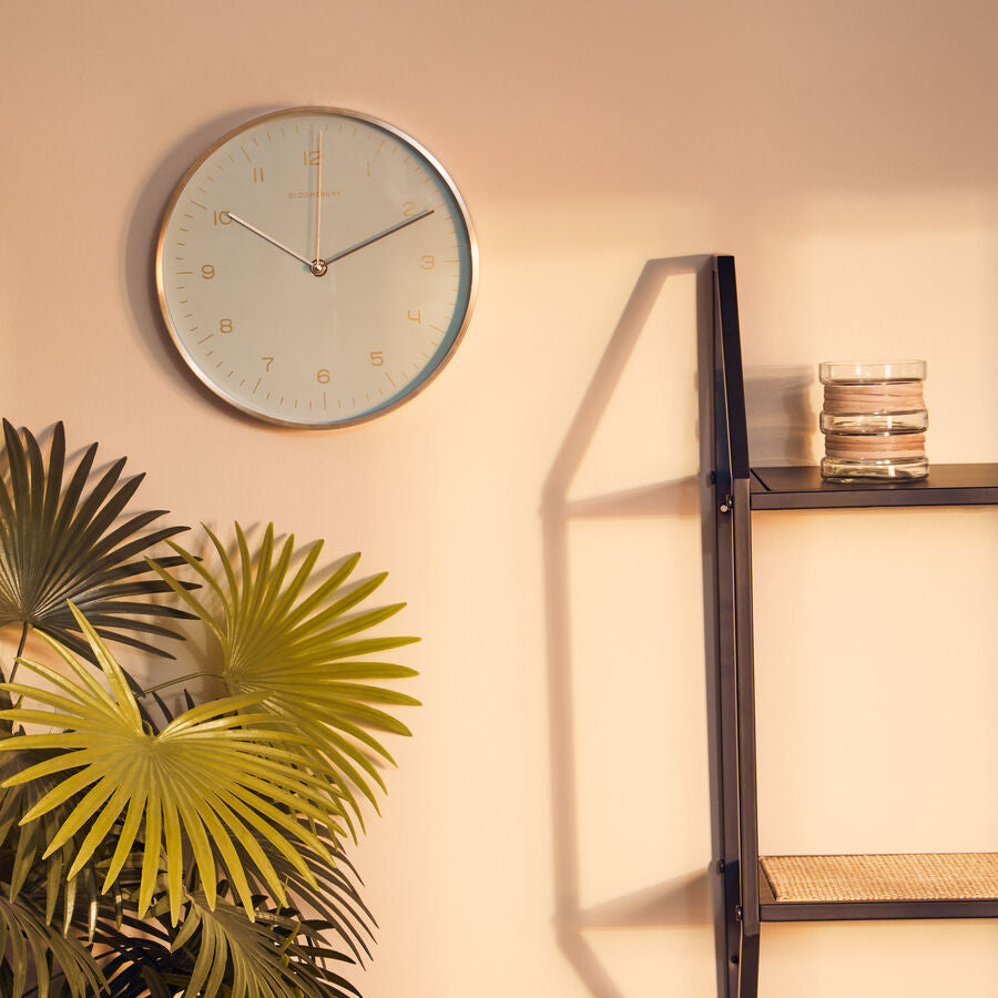 Wall clock on a beige wall with a plant and shelf in the foreground