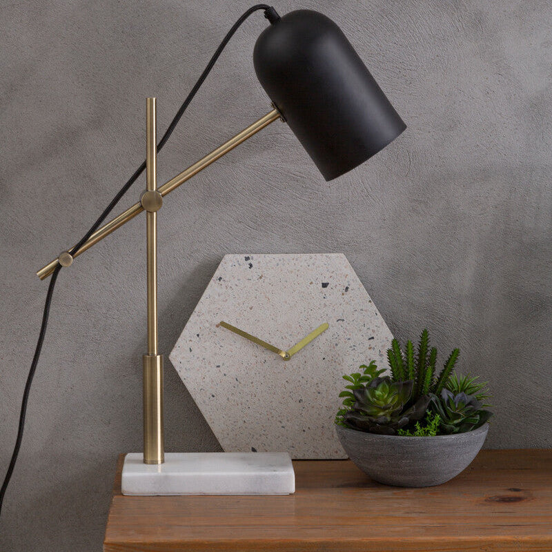 Table lamp with black shade and gold arm on a wooden table against a gray wall.