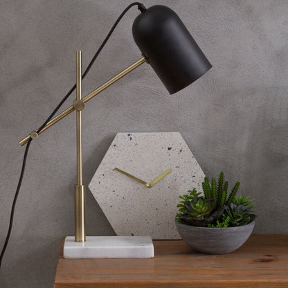 Table lamp with black shade and gold arm on a wooden table against a gray wall.