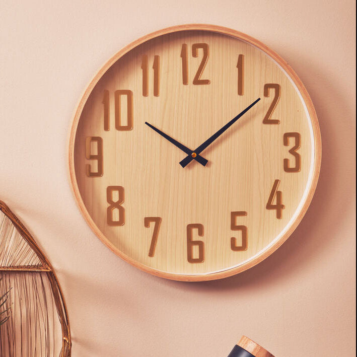 Wooden wall clock on a beige wall with a lamp and plant decoration.