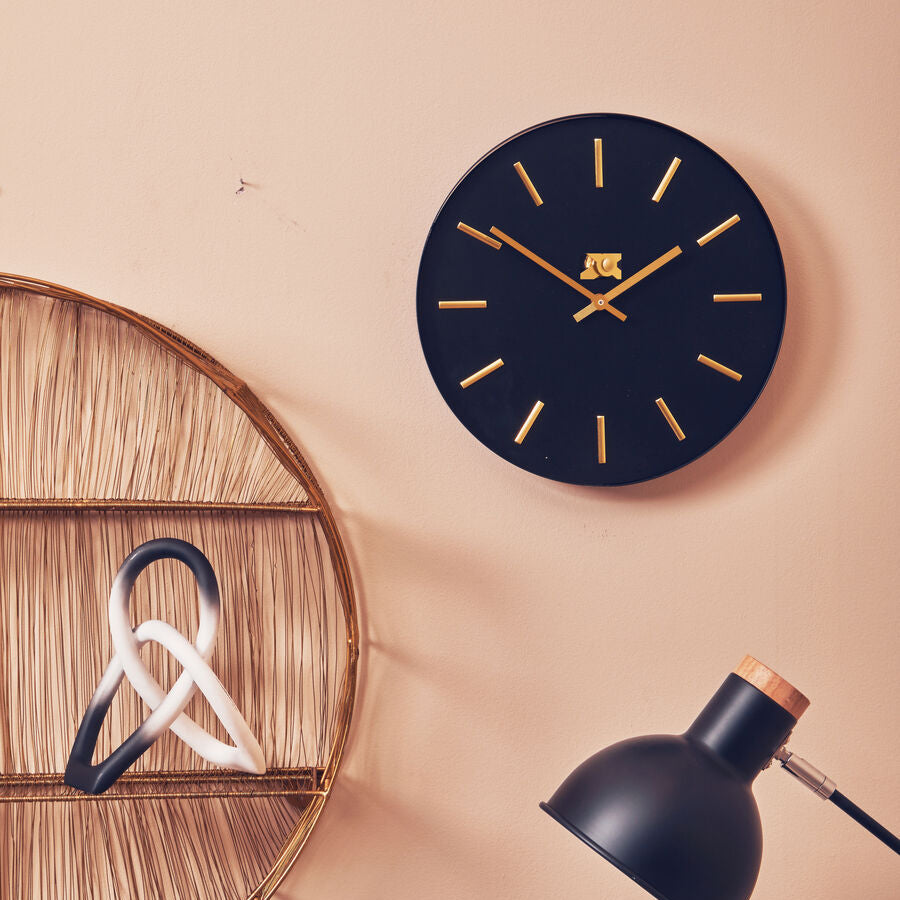Black wall clock with gold hands on a beige wall, next to a decorative shelf and lamp.