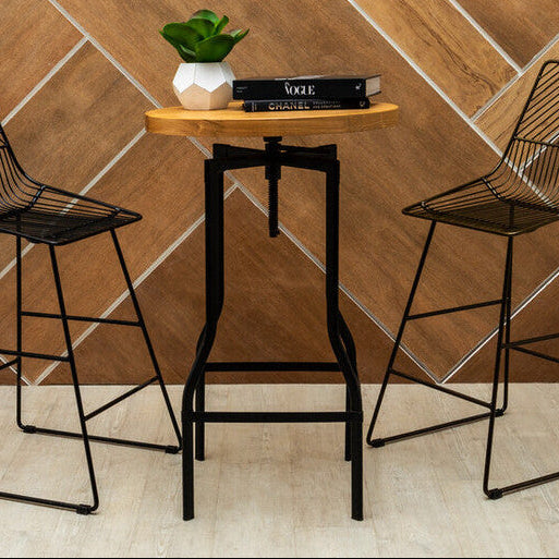 Two black wire bar stools and a small round table with a plant and books against a wooden paneled wall.