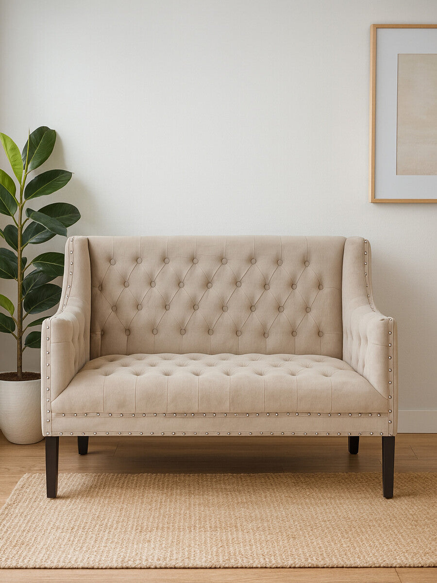 Beige tufted loveseat in a room with a plant and framed picture on the wall.