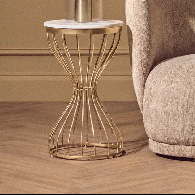 Gold side table with white lampshade next to a beige armchair against a brown paneled wall.