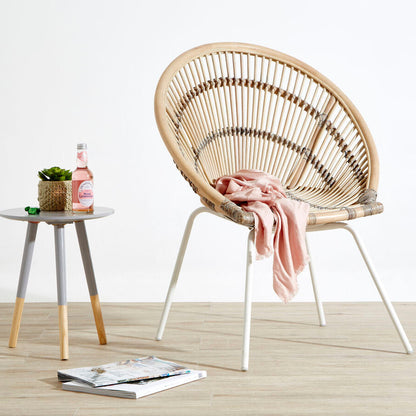Wicker chair with a pink blanket on a wooden floor, next to a small table with a bottle and plant.