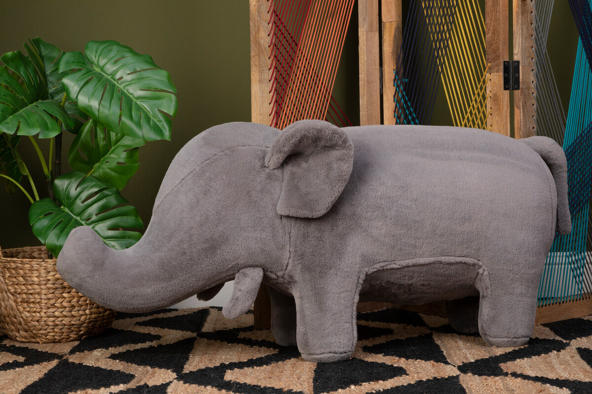 Gray elephant plush toy on a patterned rug with a plant and colorful fabric in the background