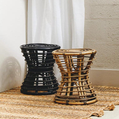 Two woven stools, one black and one natural, on a textured surface with a white curtain in the background.