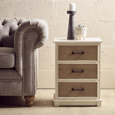 White wooden side table with three drawers next to a gray sofa against a beige wall.