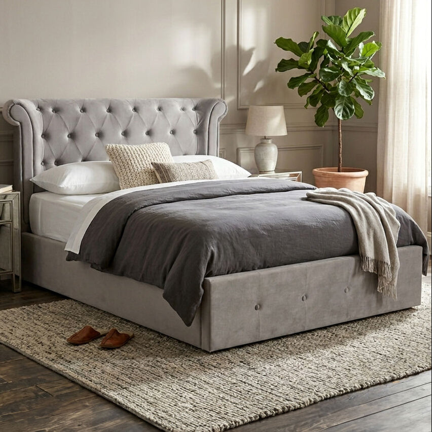 A gray upholstered bed with a tufted headboard and matching footboard, styled in a neutral bedroom with gray bedding, white pillows, a textured rug, wood flooring, bedside tables, lamps, and a potted plant.