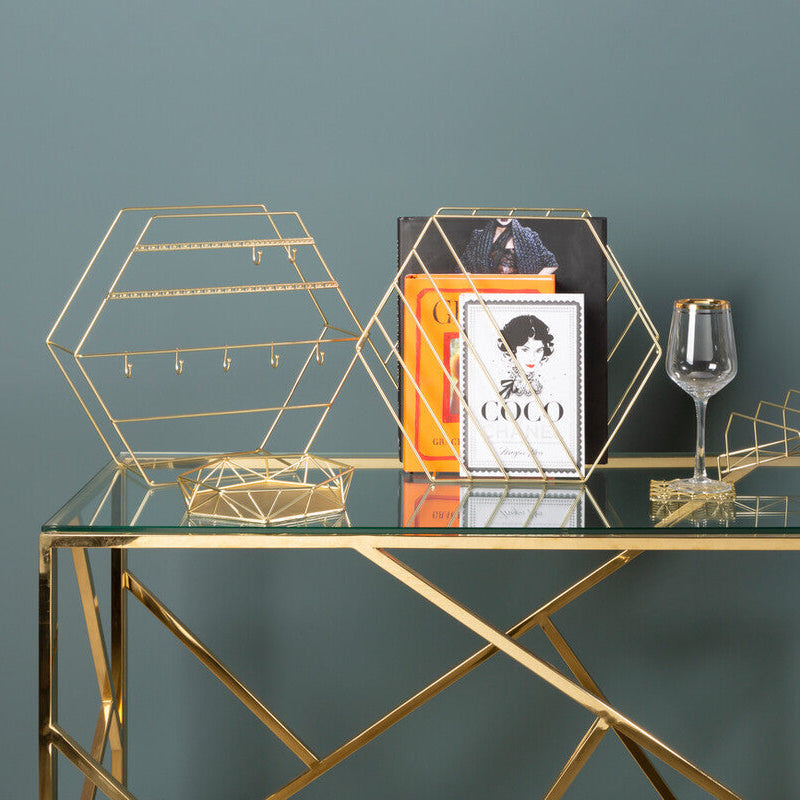 Glass table with gold geometric legs displaying decorative items against a green wall.