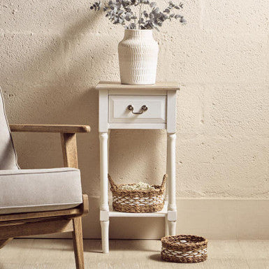 White side table with a vase of flowers and baskets, next to a wooden chair against a beige wall.