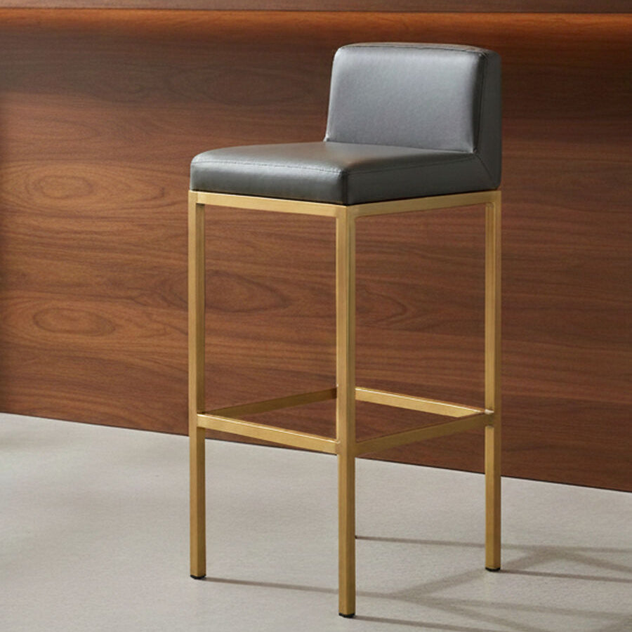 Bar stool with black cushioned seat and gold frame against a wooden wall.