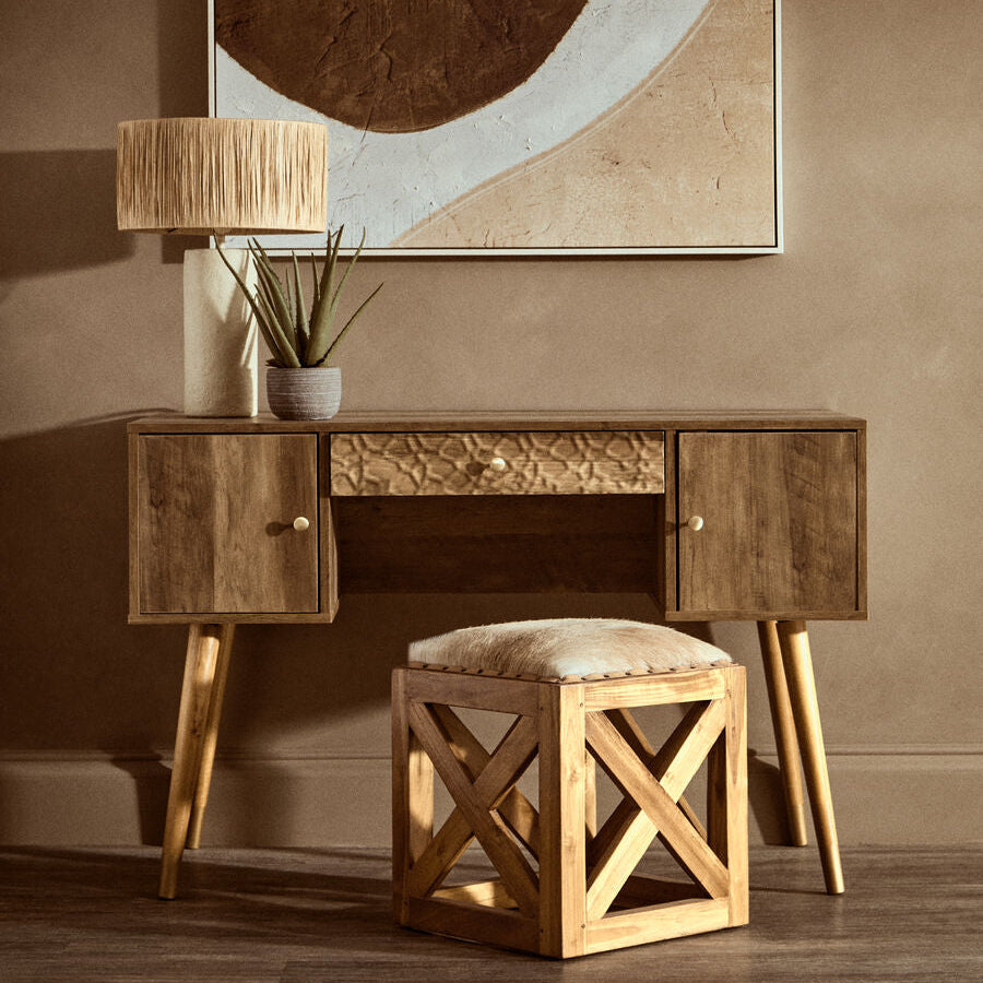 Wooden console table with a lamp, plant, and stool against a wall with abstract art.