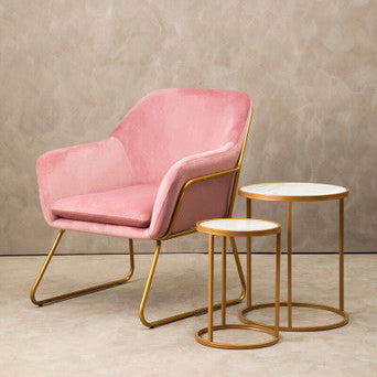 Pink armchair with gold legs and two matching side tables on a beige floor against a neutral wall.
