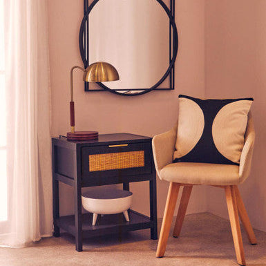 Navy side table with gold lamp, round mirror, and beige chair with black pillow in a room.