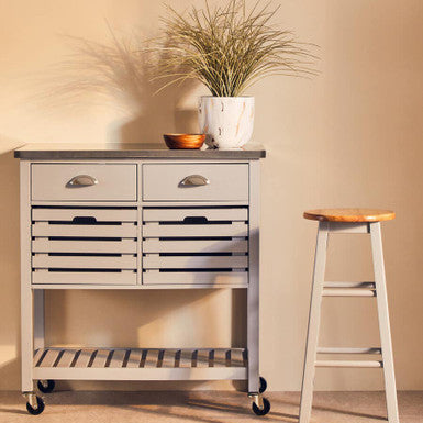 Kitchen island cart with drawers, a plant, and a stool against a beige wall.