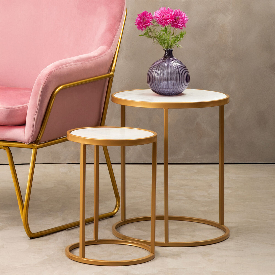 Two gold side tables with white tops next to a pink chair, one table holding a purple vase with pink flowers.