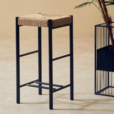 Bar stool with woven seat and black metal frame against a beige wall with a plant.