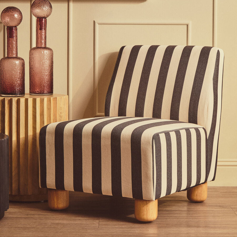 A modern accent chair upholstered in bold black and cream vertical stripes, styled in a warm, beige-toned room next to decorative glass bottles.
