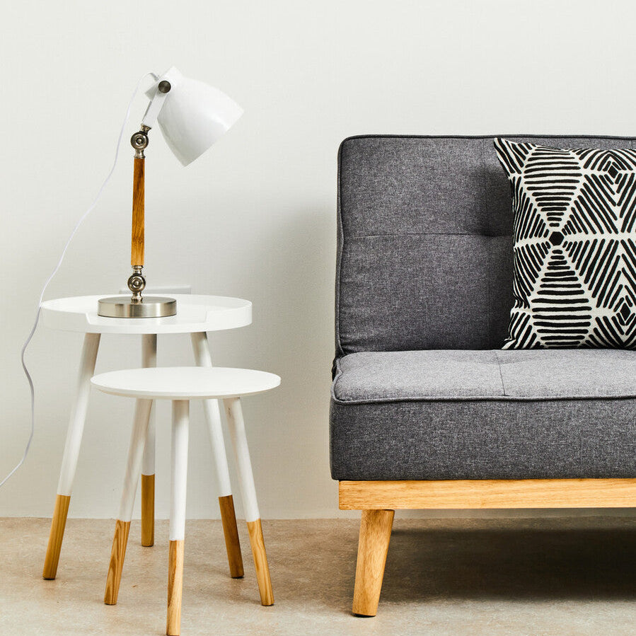 Gray sofa with a patterned pillow, white side table with lamp, and small round stool in a minimalistic room.