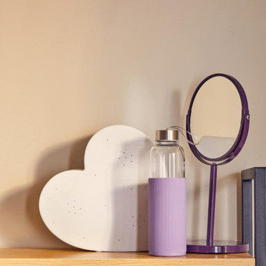 Heart-shaped object, clear bottle with purple sleeve, and round mirror on a wooden shelf against a beige wall.
