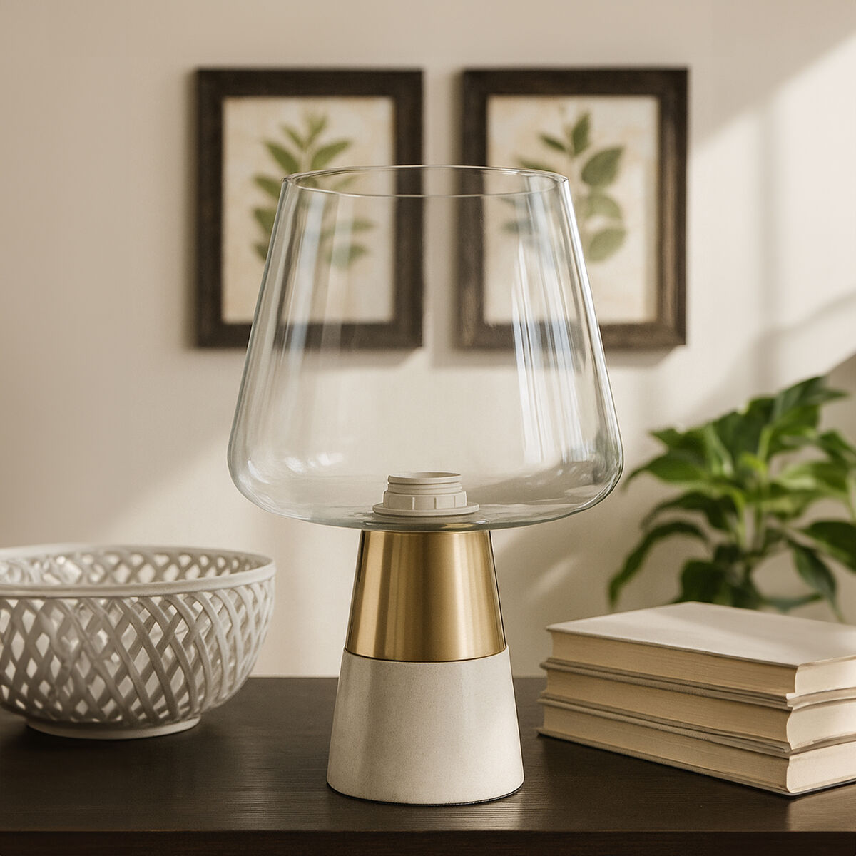 Clear glass table lamp with gold and white base on a wooden surface, with books and decorative items in the background.