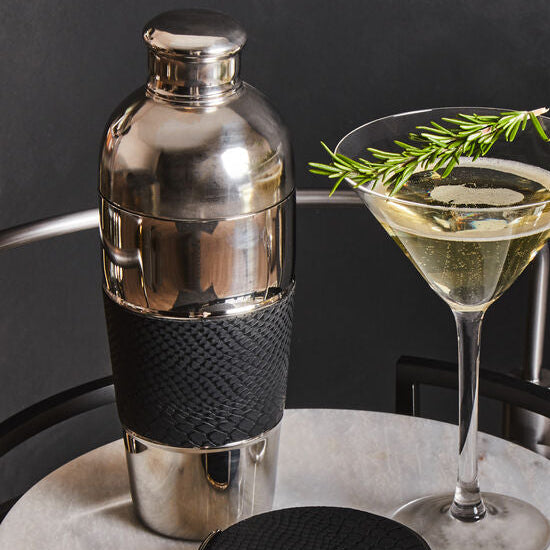 Two cocktails in martini glasses with a shaker on a round black tray against a dark background.