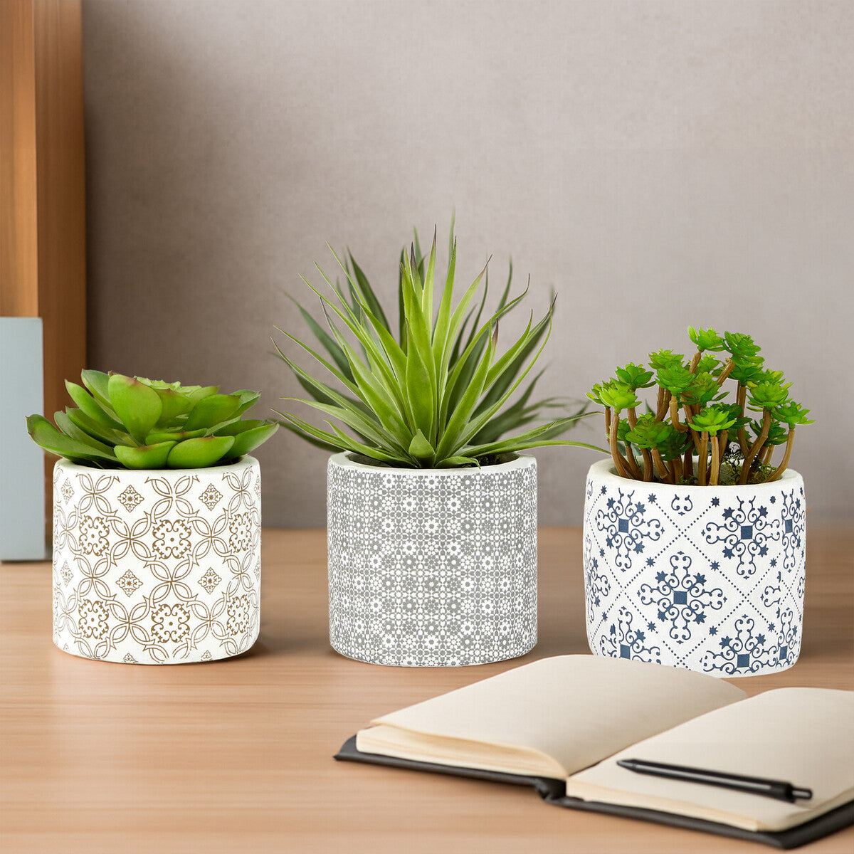 Three patterned planters with plants on a wooden surface next to an open notebook.