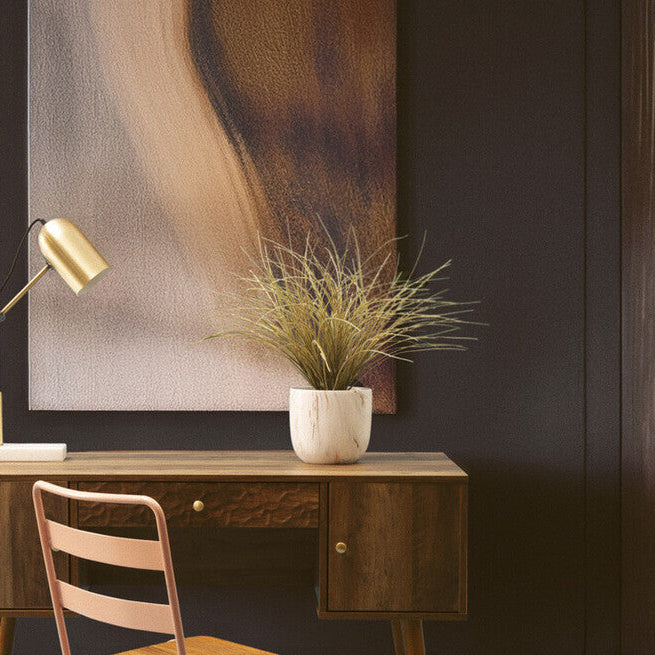 Wooden desk with chair, lamp, and plant against a dark wall with abstract painting