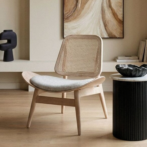 A modern minimalist living room featuring a light wood and woven cane lounge chair with a light gray cushion, positioned next to a black ribbed side table. A large abstract art print with brown and beige marbled swirls hangs on the beige wall behind the chair.