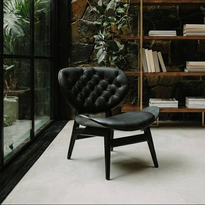 A black tufted leather armchair with dark wooden legs is placed in a modern interior with a stone wall, shelving, books, and large windows with green plants.