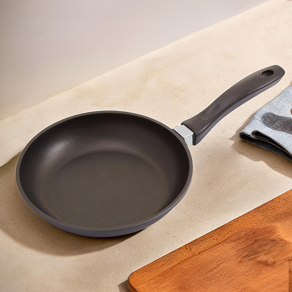 Black frying pan on a beige surface with a wooden cutting board and gray towel in the background