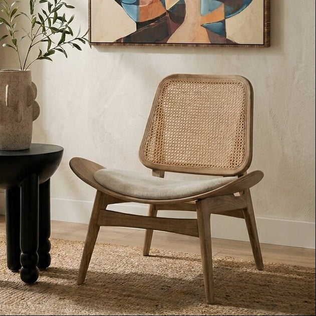 An abstract framed canvas print with geometric shapes in blue, beige, brown, and black, displayed on a textured beige wall in a styled room setting with a wooden chair, a black side table, and a potted plant.