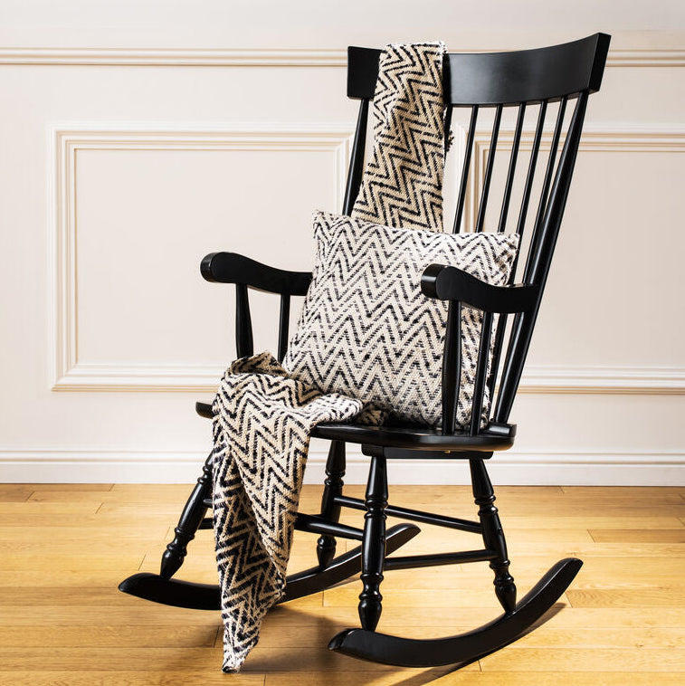 Black rocking chair with patterned cushions and blanket on a wooden floor against a white wall.