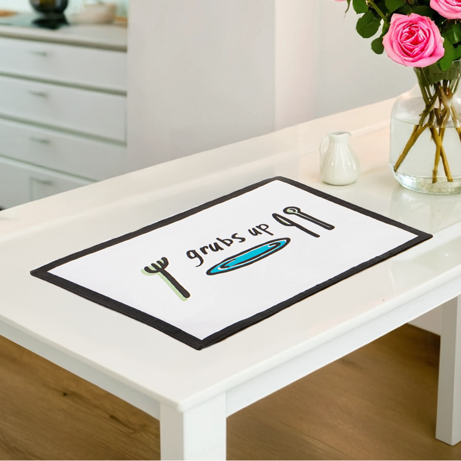Table with a decorative placemat featuring cutlery designs, next to a vase of pink roses in a kitchen setting.