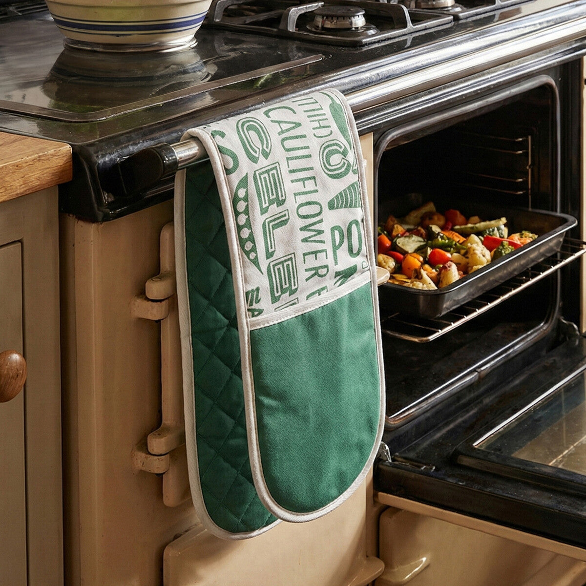 Green oven mitt hanging on a stove with an open oven showing roasted vegetables.