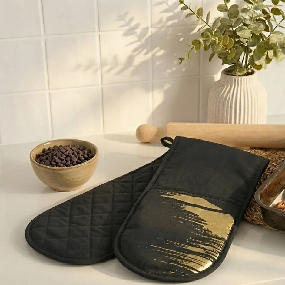 Black oven mitt with gold interior on a kitchen counter with cookies and coffee beans.