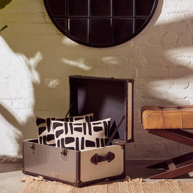 Open suitcase with patterned pillows on a textured rug, against a white brick wall with a round mirror.