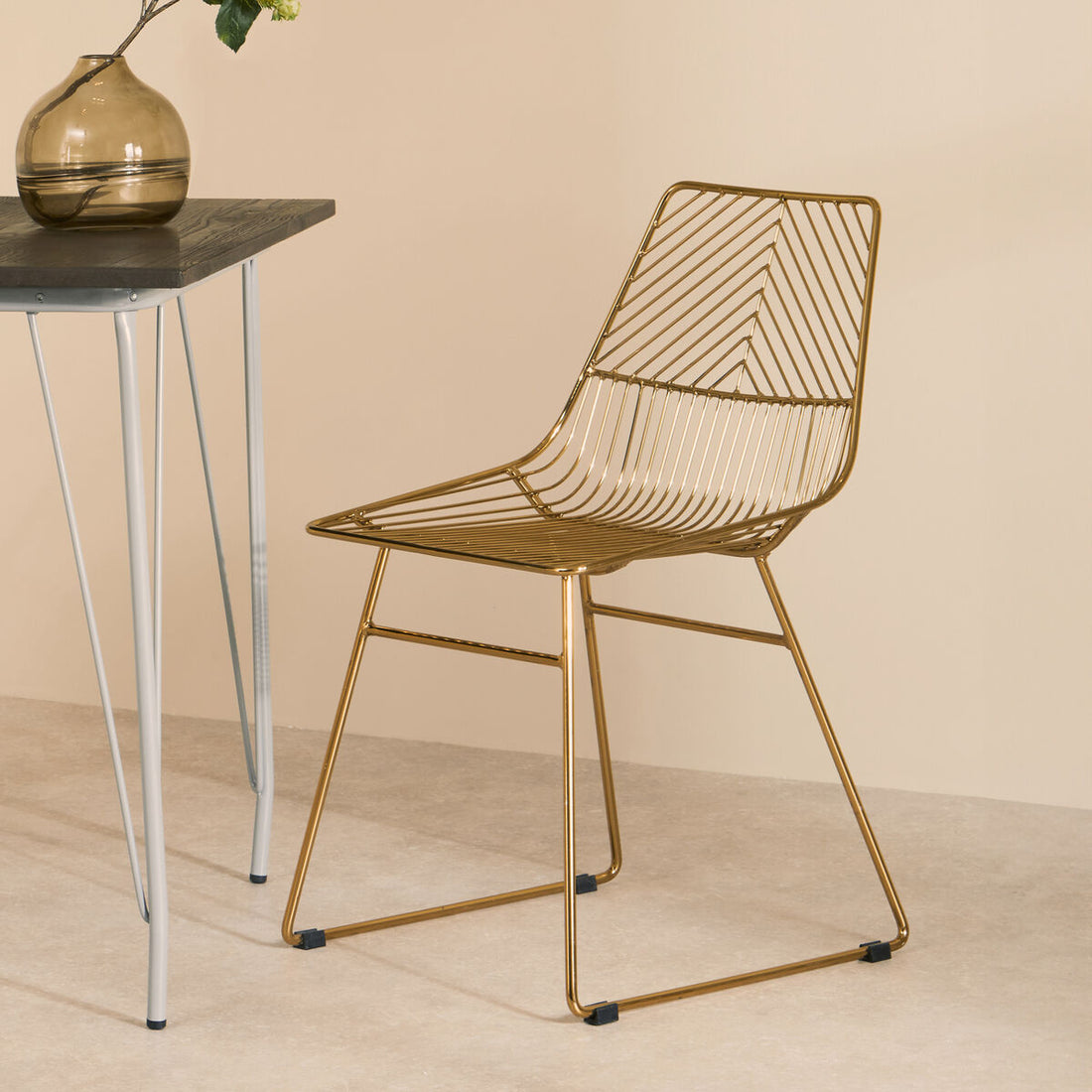Gold wire chair next to a small table with a vase on a beige background
