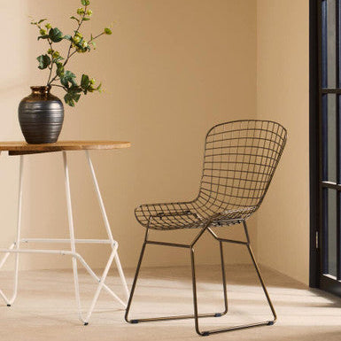 Wire chair next to a wooden table with a vase and plant against a beige wall.