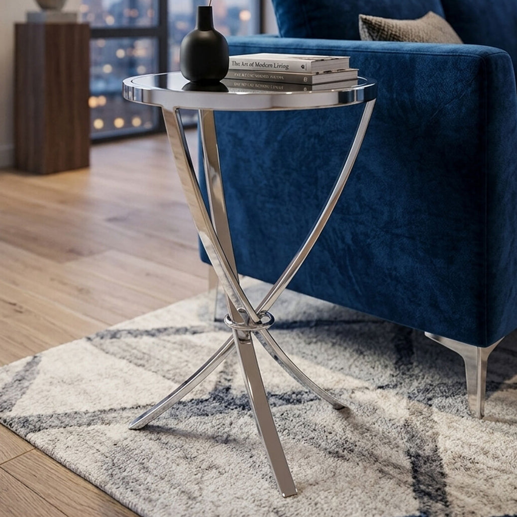 A modern round side table with a silver chrome metal frame and a clear glass top, styled in a living room next to a blue velvet sofa.