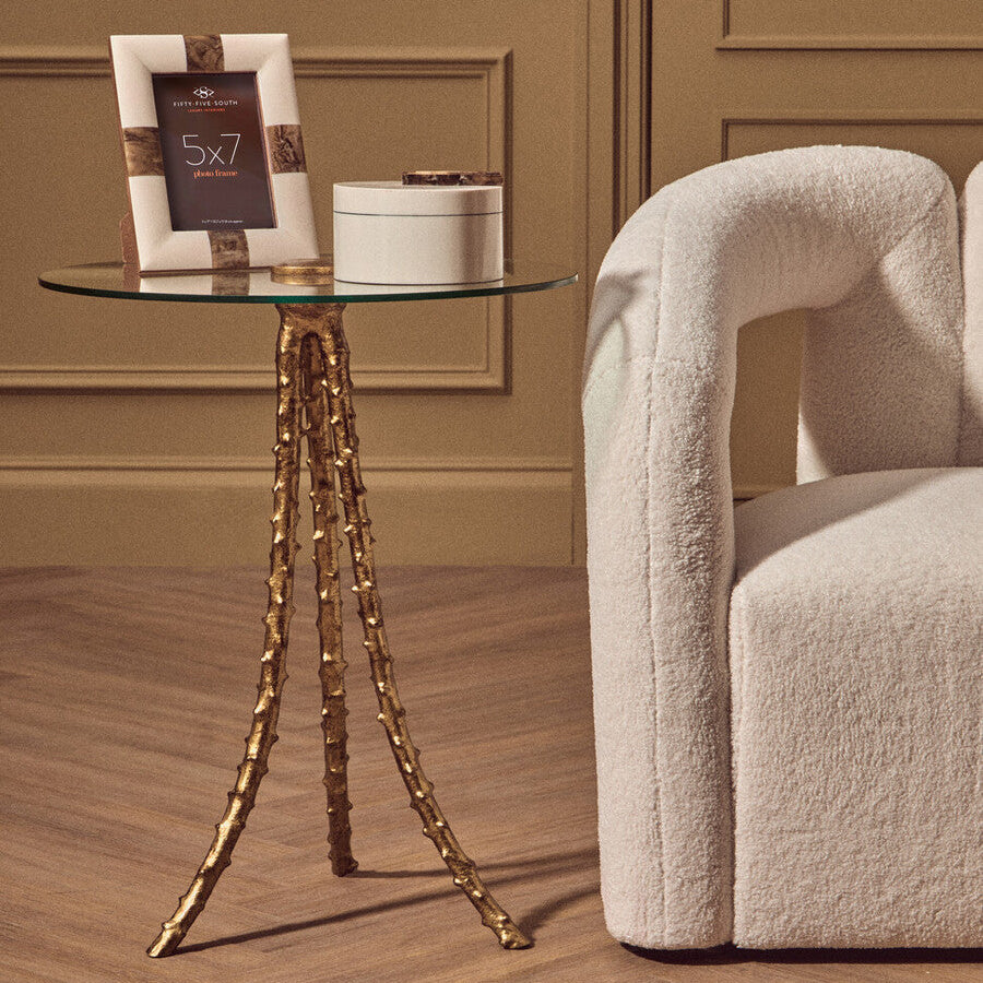 Glass side table with gold legs next to a textured beige armchair in a room with wooden flooring and paneled walls.