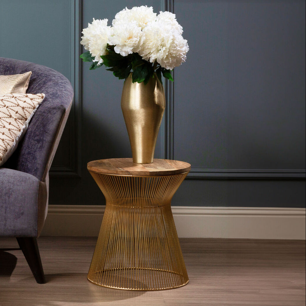 A gold wire side table holding a matching gold vase filled with white flowers, styled next to a purple armchair in a room with dark paneled walls and wood flooring.