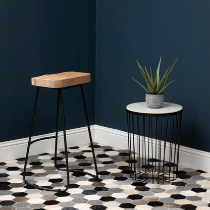 Stool with wooden seat and black metal legs next to a small round table with a plant on a hexagonal patterned rug.